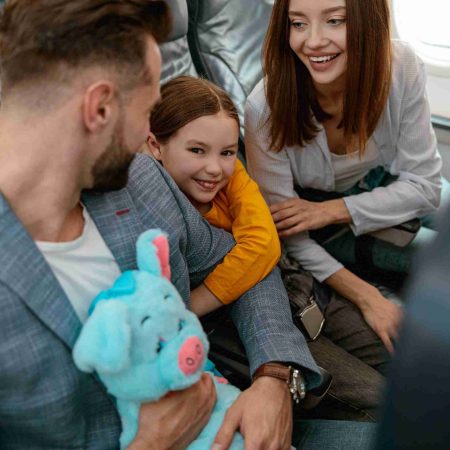 Cheerful little girl holding father hand and smiling while sitting next to mother in passenger aircraft